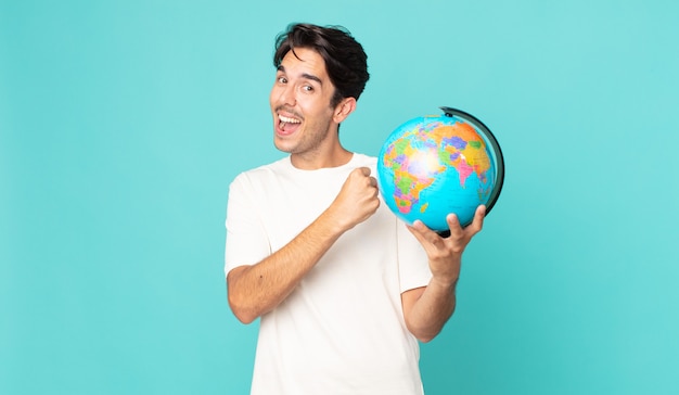 Young hispanic man feeling happy and facing a challenge or celebrating and holding a world globe map