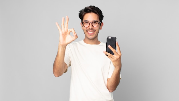 Young hispanic man feeling happy, showing approval with okay gesture and holding a smartphone