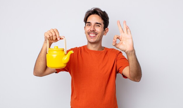 young hispanic man feeling happy, showing approval with okay gesture. teapot concept