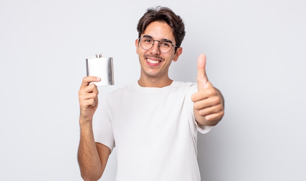 Young hispanic man feeling proud,smiling positively with thumbs up. alcohol flask concept