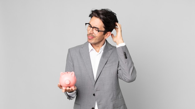 Young hispanic man feeling puzzled and confused, scratching head and holding a piggy bank