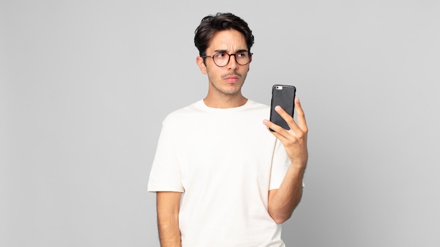 Young hispanic man feeling sad, upset or angry and looking to the side and holding a smartphone