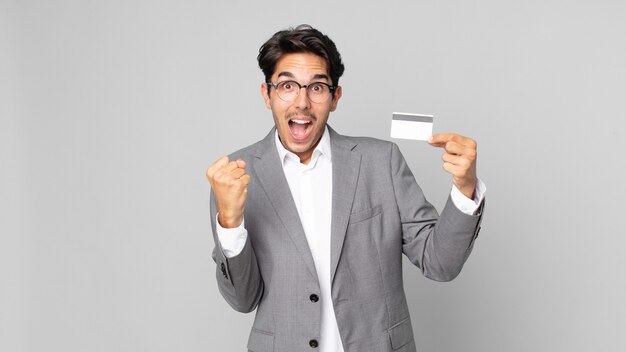 Young hispanic man feeling shocked,laughing and celebrating success and holding a credit card