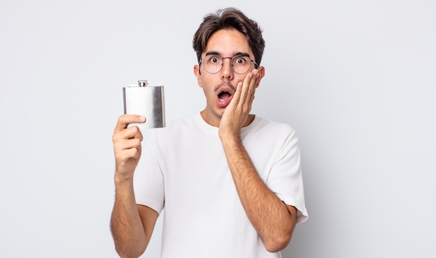 Young hispanic man feeling shocked and scared. alcohol flask concept