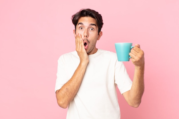 Young hispanic man feeling shocked and scared and holding a coffee mug