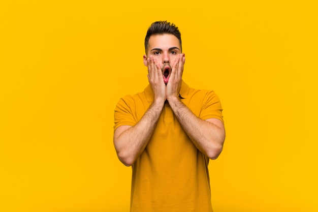 Young hispanic man feeling shocked and scared, looking terrified with open mouth and hands on cheeks against orange wall