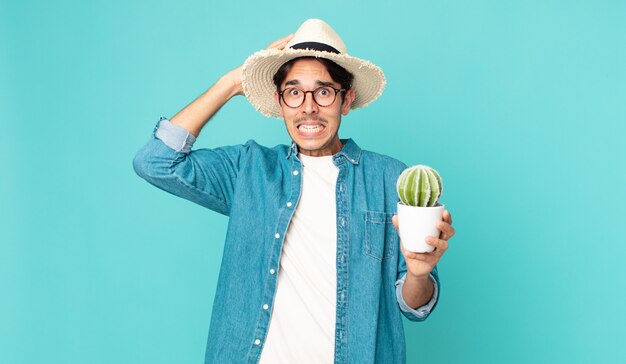 young hispanic man feeling stressed, anxious or scared, with hands on head and holding a cactus