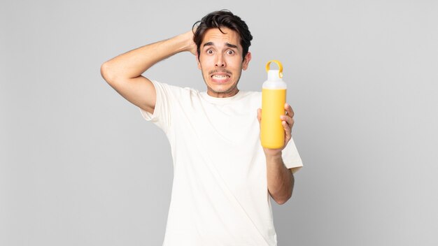 young hispanic man feeling stressed, anxious or scared, with hands on head with a coffee thermos