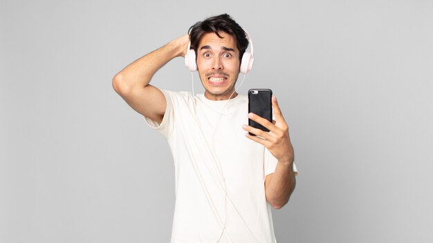 Young hispanic man feeling stressed, anxious or scared, with hands on head with headphones and smartphone