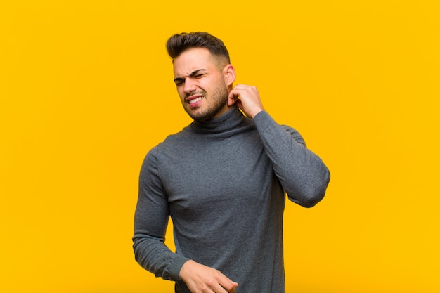 Young hispanic man feeling stressed, frustrated and tired, rubbing painful neck, with a worried, troubled look against orange wall