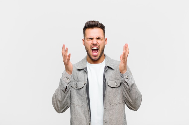 Young hispanic man furiously screaming, feeling stressed and annoyed with hands up in the air saying why me against gray background