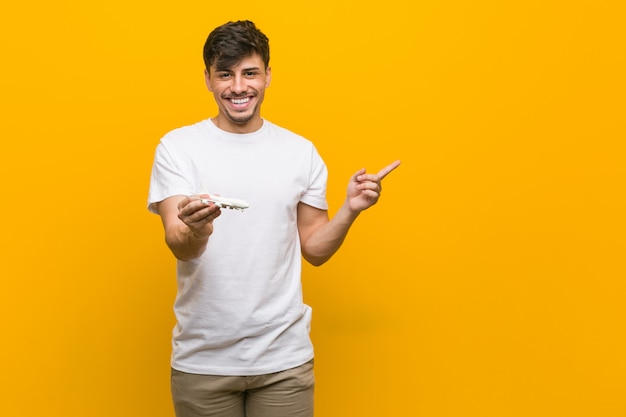 Young hispanic man holding an airplane icon smiling cheerfully pointing with forefinger away.