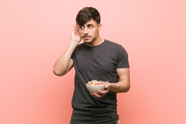 Young hispanic man holding a cereal bowl trying to listening a gossip.