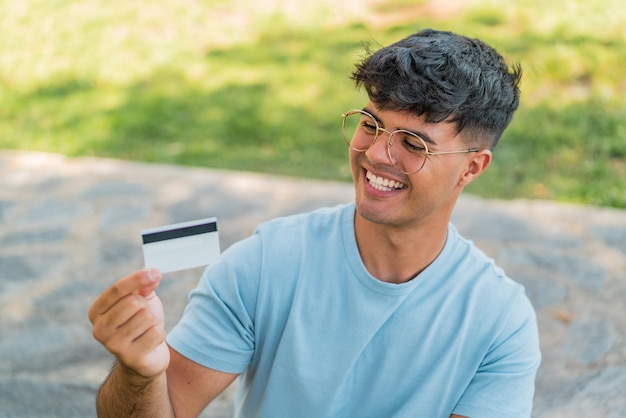 Young hispanic man holding a credit card at outdoors with happy expression