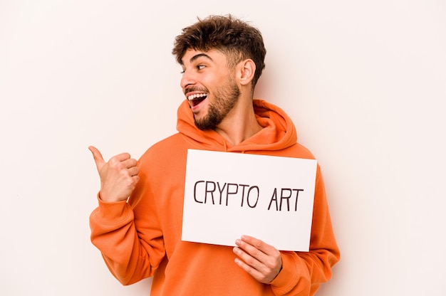Молодой латиноамериканец, держащий плакат с крипто-искусством, изолированный на белом фоне, показывает большим пальцем в сторону, смеясь и беззаботно