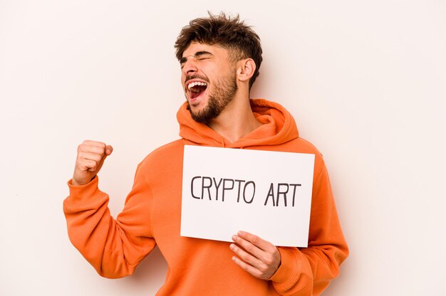 Young hispanic man holding a crypto art placard isolated on white background raising fist after a victory winner concept