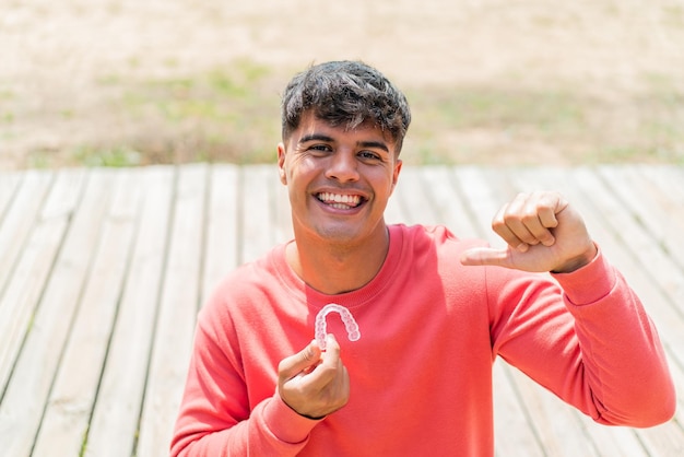 Photo young hispanic man holding invisible braces at outdoors proud and selfsatisfied