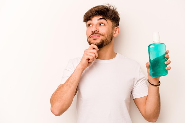 Young hispanic man holding mouthwash isolated on white background looking sideways with doubtful and skeptical expression