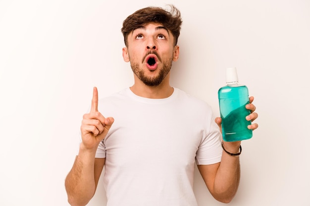 Young hispanic man holding mouthwash isolated on white background pointing upside with opened mouth