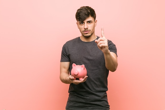 Young hispanic man holding piggy bank showing number one with finger.