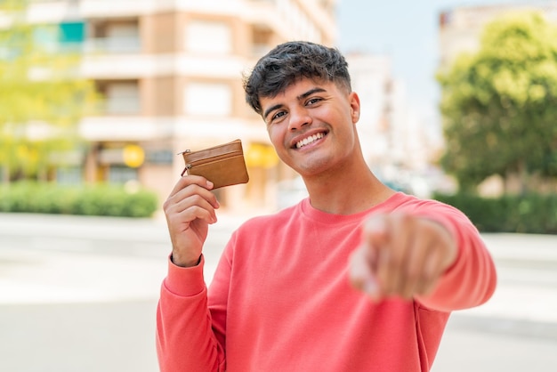 Young hispanic man holding a wallet at outdoors points finger at you with a confident expression