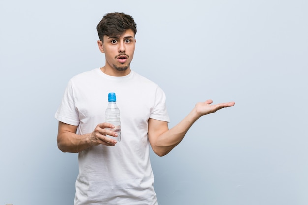 Young hispanic man holding a water bottle impressed holding blank space on palm.