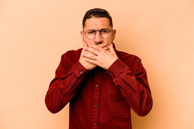 Young hispanic man isolated on beige background covering mouth with hands looking worried