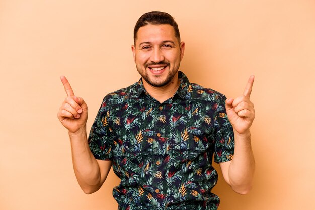 Young hispanic man isolated on beige background indicates with both fore fingers up showing a blank space