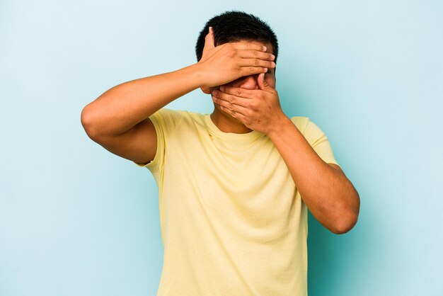 Young hispanic man isolated on blue background blink at the camera through fingers embarrassed covering face