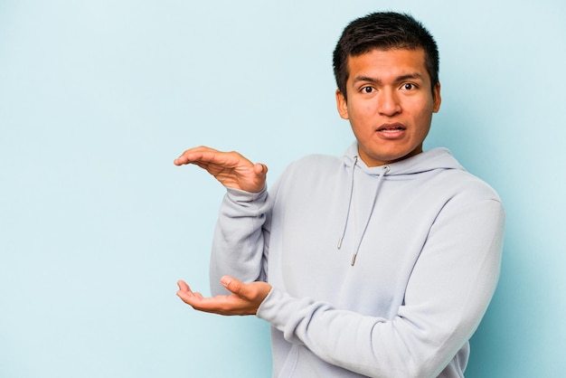 Young hispanic man isolated on blue background shocked and amazed holding a copy space between hands