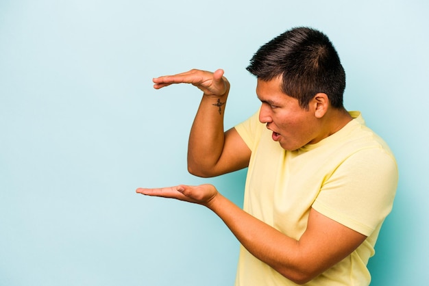 Young hispanic man isolated on blue background shocked and amazed holding a copy space between hands
