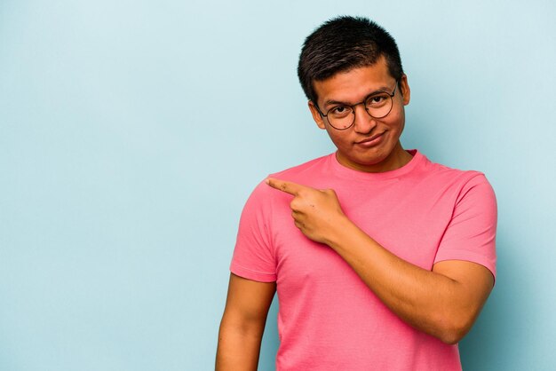 Young hispanic man isolated on blue background smiling and pointing aside showing something at blank space