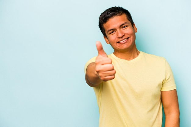 Молодой латиноамериканец на синем фоне улыбается и поднимает большой палец вверх