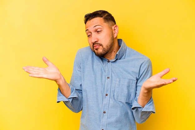 Young hispanic man isolated on yellow background confused and doubtful shrugging shoulders to hold a copy space