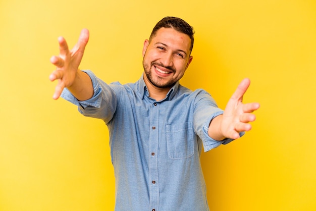 Young hispanic man isolated on yellow background feels confident giving a hug to the camera