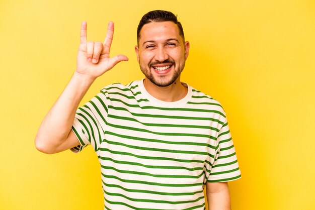 Young hispanic man isolated on yellow background showing a horns gesture as a revolution concept