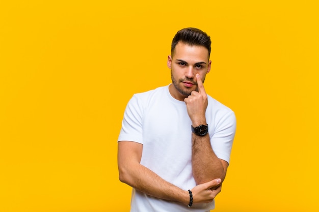 Young hispanic man keeping an eye on you, not trusting, watching and staying alert and vigilant against orange wall