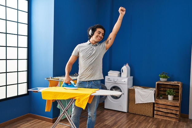 Young hispanic man listening to music ironing clothes at laundry room