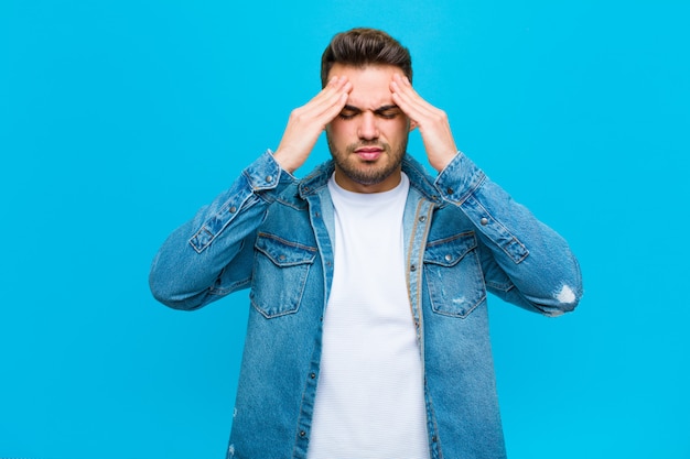 Young hispanic man looking concentrated, thoughtful and inspired, brainstorming and imagining with hands on forehead against blue wall