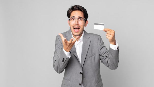 Young hispanic man looking desperate, frustrated and stressed and holding a credit card