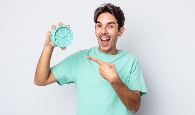 young hispanic man looking excited and surprised pointing to the side. alarm clock concept