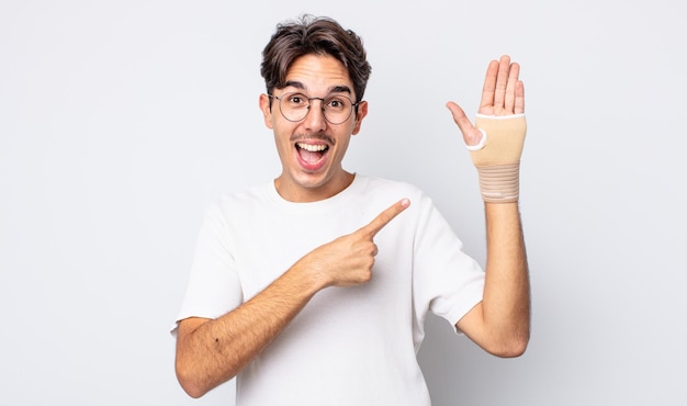 Young hispanic man looking excited and surprised pointing to the side. hand bandage concept