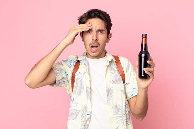 Young hispanic man looking happy, astonished and surprised and holding a bottle of beer