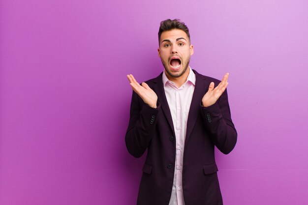 Young hispanic man looking happy and excited, shocked with an unexpected surprise with both hands open next to face