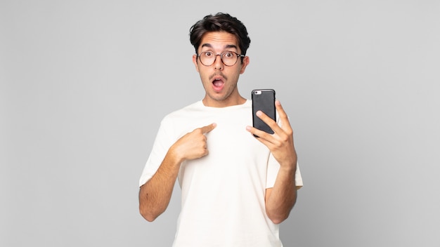 Young hispanic man looking shocked and surprised with mouth wide open, pointing to self and holding a smartphone