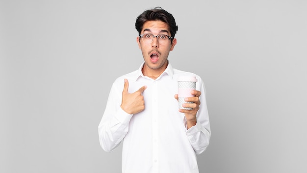 Young hispanic man looking shocked and surprised with mouth wide open, pointing to self and holding a take away coffee