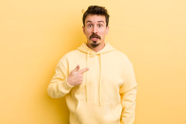 Young hispanic man looking shocked and surprised with mouth wide open pointing to self