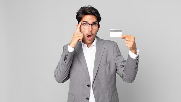 young hispanic man looking surprised, realizing a new thought, idea or concept and holding a credit card