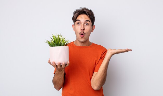 Young hispanic man looking surprised and shocked, with jaw dropped holding an object. decorative plant concept