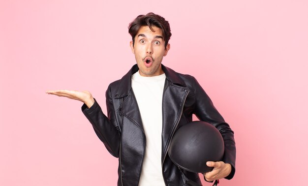 Young hispanic man looking surprised and shocked, with jaw dropped holding an object. motorbike rider concept